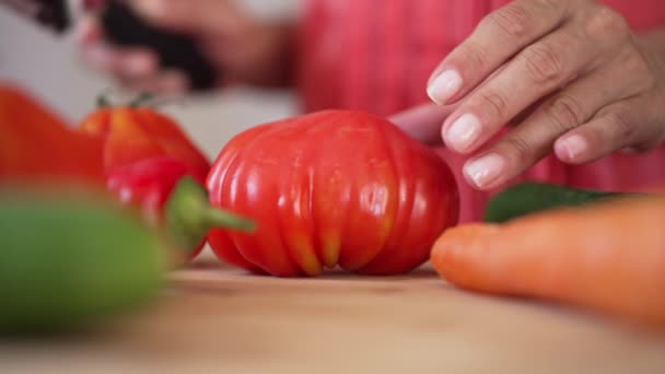 Close-up cutting red ripe tomato with knife on cutting board in slow motion. Unrecognizable Caucasian woman turning halves to camera standing indoors in kitchen. Dinner and food concept. — ストック動画