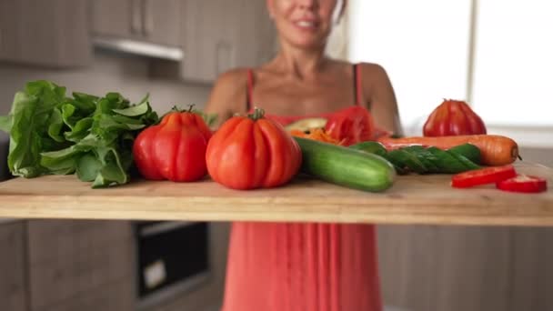 Close-up healthful organic vegetables on wooden cutting board with blurred unrecognizable Caucasian woman smiling at background. Happy housewife bragging salad ingredients indoors. — Vídeo de Stock