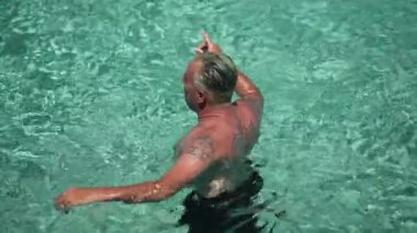 High angle view of tattooed tanned man standing in turquoise Mediterranean sea in sunshine. Caucasian male tourist enjoying summer vacations outdoors swimming in ocean lagoon. Slow motion.