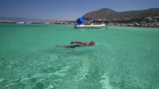 Seitenansicht selbstbewusst tätowierter, kaukasisch gebräunter Mann, der in Zeitlupe im türkisfarbenen Mittelmeer schwimmt. Yachten in der Lagune im Hintergrund. Urlaubs- und Erholungskonzept. — Stockvideo