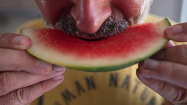Close-up red watermelon slice with unrecognizable bearded man eating delicious juicy berry in slow motion. Caucasian man enjoying taste of sweet dessert on summer day indoors. — Stok video