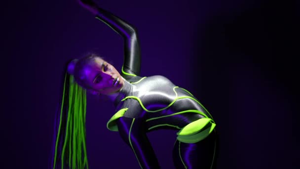 Flexible slim woman bending back raising leg standing on cube in ultraviolet light. Caucasian young gymnastic performing modern dance in fluorescent neon looking at camera. — Stock video