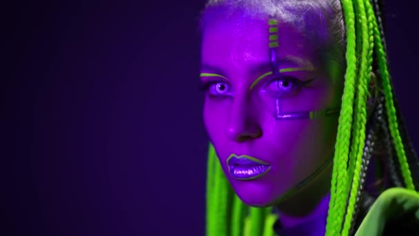 Headshot of slim woman with futuristic makeup looking at camera as strobe lights turning on. Live camera zoom out as flexible performer singing dancing bending on cube in ultraviolet. — Wideo stockowe