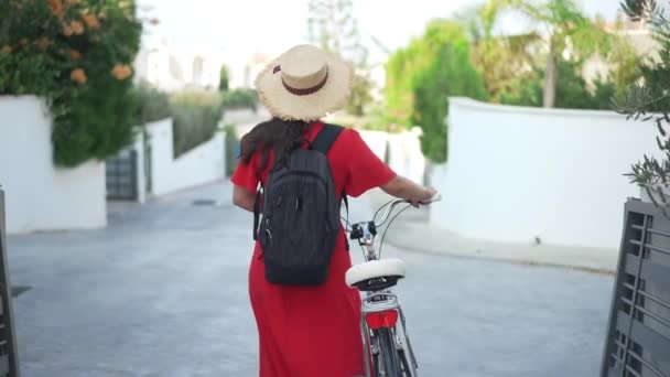Вид сзади молодая женщина-велосипедистка с велосипедом, идущая по улице в замедленном режиме. Живая камера следит за уверенной кавказской женщиной в красном платье и соломенной шляпе, прогуливающейся на велосипеде по улице Цитруса. — стоковое видео