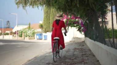 Kıbrıs caddesinde yavaş çekimde bisiklete binen kırmızı elbiseli beyaz bir kadın. Geniş açılı, mutlu bir turist dışarıda güneşli bir yaz tatili geçiriyor. Yaşam tarzı ve neşe kavramı.