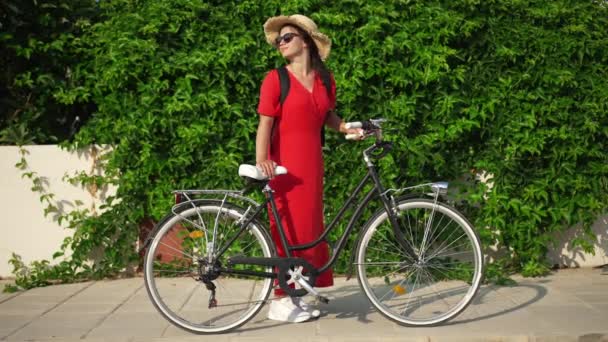 自転車で屋外に立って赤いドレスで幸せな自信に満ちた豪華な若い女性の広いショットの肖像画は、スローモーションで残し周りを見回します。キプロスで夏の日を楽しむ白人観光客. — ストック動画