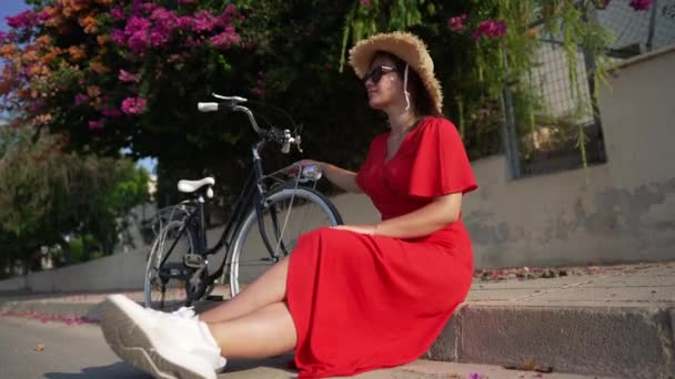 Joven mujer caucásica en vestido rojo sentada en la calle con bicicleta mirando a su alrededor admirando la naturaleza de Chipre. Retrato de turista confiado descansando bajo el sol al aire libre en el día de verano. Movimiento lento. — Vídeo de stock