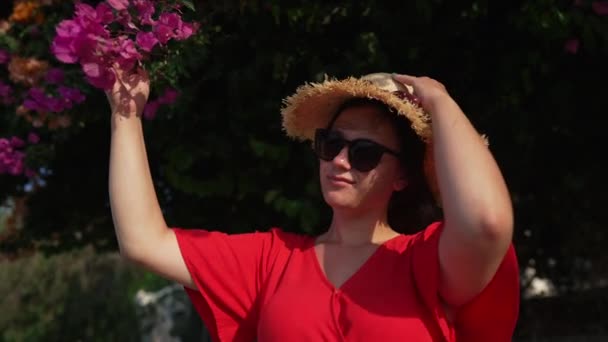 Portrait of young happy woman putting on straw hat in slow motion holding flower on tree outdoors. Satisfied Caucasian female tourist enjoying vacations on Cyprus. Lifestyle and leisure. — Stockvideo