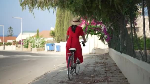 Back view young Caucasian woman in red dress riding bicycle in slow motion on Cyprus street. Wide shot happy tourist enjoying summer vacations in sunbeam outdoors. Lifestyle and joy concept. — Vídeos de Stock