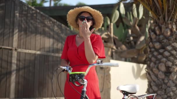 Koketa Kavkazská mladá žena gestikuluje vzduchový polibek a usmívá se na kameru stojící venku na kole. Přední pohled portrét šťastného bezstarostného tisíciletého turisty pózujícího ve zpomaleném filmu ve slunečním světle. — Stock video
