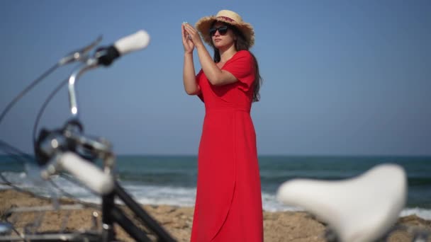 Confident young woman dancing in slow motion standing on Mediterranean sea beach smiling with blurred bike at front. Portrait of happy Caucasian gorgeous tourist enjoying vacations. — ストック動画