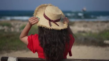 Arkadan bakıldığında, tanınmayan, hasır şapka takan, bankta oturan Akdeniz 'e hayran genç bir kadın. Beyaz kadın milenyum turisti Kıbrıs 'ta açık havada yaz gününün tadını çıkarıyor. Yavaş çekim.