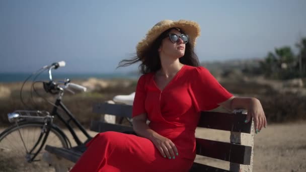 Розслаблена усміхнена жінка у сонцезахисних окулярах, що сидять на лавці з велосипедом на вулиці. Портрет щасливого безтурботного кавказького велосипедиста насолоджується літньою відпусткою на узбережжі Середземного моря.. — стокове відео