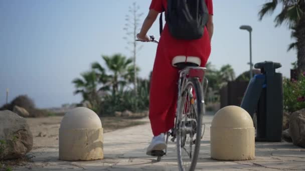Beautiful Cyprus landscape with young woman in red dress passing on bike riding in slow motion away. Back view slim confident Caucasian female tourist biking outdoors on summer day. Leisure and joy. — Stok video