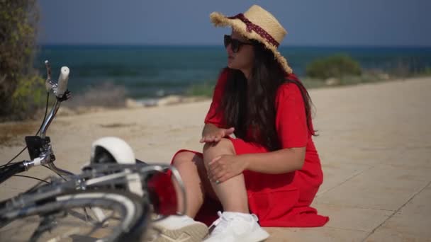 Sad young Caucasian woman rubbing knee fell from bicycle outdoors on sunny summer day. Portrait of upset beautiful bicyclist sitting at background of Mediterranean sea with injured leg. — 图库视频影像