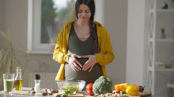 행복하게 임신 한 젊은 여성이 웃고 있는 카메라를 보면서 심장 모양을 꾸미고 있는 모습. 앞을 보고 건강에 좋은 샐러드 재료와 테이블에 포즈를 취하는 밝은 캅카스 천 년 대. — 비디오