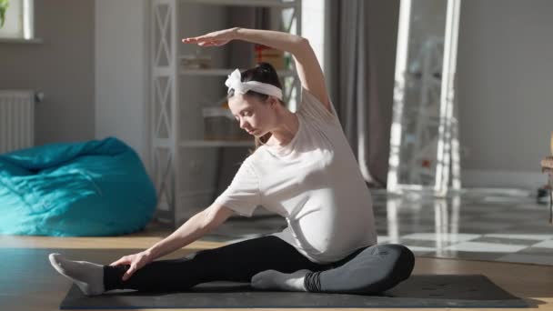 Flexible young pregnant woman bending aside sitting on exercise mat on sunny morning at home. Wide shot portrait of fit athletic confident Caucasian sportswoman training indoors. Pregnancy and sport. — стоковое видео