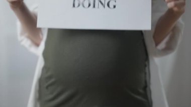 Close-up pregnant belly with female hands lowering Think before doing placard. Unrecognizable young Caucasian woman calling for consciousness. Pregnancy and lifestyle.