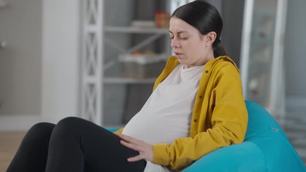 Side view pregnant woman having contractions calling hospital talking on phone. Young Caucasian worried expectant sitting on bag chair indoors. Childbirth concept. — ストック動画