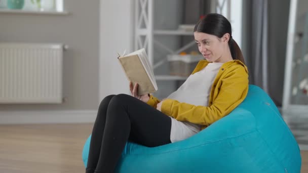Side view smiling pregnant woman reading book stroking belly sitting on bag chair indoors. Portrait of happy Caucasian young expectant enjoying hobby with unborn baby at home. — ストック動画