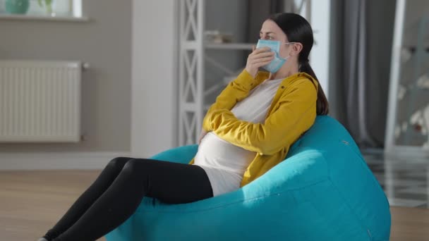 Unwell Caucasian pregnant woman in coronavirus face mask coughing sitting at home indoors. Side view portrait of unhealthy young ill expectant. Covid-19 pandemic and pregnancy concept. — Stok video