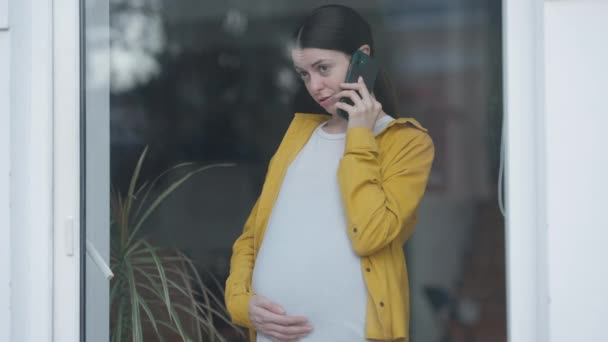 Krásná pozitivní těhotná žena mluví po telefonu, stojí u skleněných dveří a dívá se ven. Portrét šťastné sebevědomé kavkazské očekávání těší volný čas doma. — Stock video
