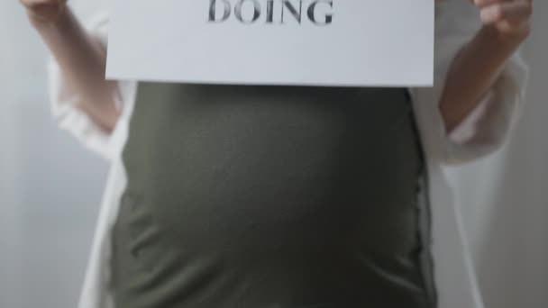 Крупным планом беременный живот с опущенными женскими руками подумайте, прежде чем делать плакат. Неузнаваемая молодая белая женщина, призывающая к сознанию. Беременность и образ жизни. — стоковое видео