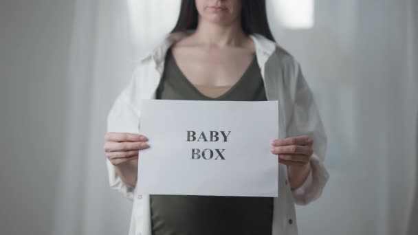 Young unrecognizable pregnant woman stretching Baby box placard to camera standing indoors. Concept of pregnancy and maternity leave. — Αρχείο Βίντεο