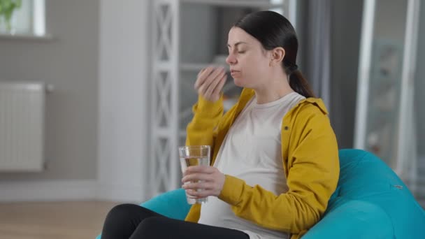 Side view pregnant woman taking in pills sitting on bag chair at home indoors. Portrait of Caucasian young expectant taking care of health. Pregnancy and medicine concept. — ストック動画