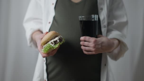 Pregnant woman with unhealthy sandwich and soda water standing indoors. Front view unrecognizable Caucasian expectant eating drinking harmful junk food. — Vídeo de Stock