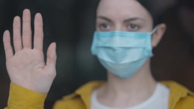 Close-up female hand on glass with blurred sad woman on coronavirus face mask at background. Upset Caucasian young ill lady standing indoors on Covid-19 pandemic quarantine looking out window.