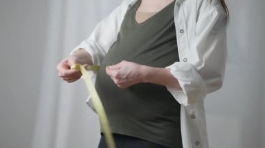 Unrecognizable pregnant woman measuring waist and belly standing indoors at home. Caucasian young slim expectant controls body weight and fetus. Health care and pregnancy.