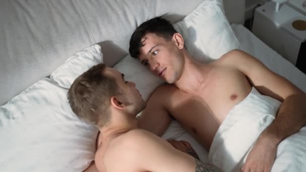 Счастливая расслабленная любящая гей пара, лежащая в удобной кровати и улыбающаяся по утрам. Позитивные кавказские юноши обнимаются дома в спальне, болтая. Концепция отношений и радости. — стоковое видео