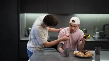 Genç eşcinsel adam evde mutfakta dikilen partnerin tişörtünü eleştiriyor. Kafkasyalı LGBTQ 'lu birinin sabah evde erkek arkadaşıyla konuşmasının yan görüntüsü..