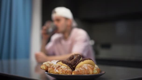 Primer plano deliciosas rosquillas dulces en el plato con borrosa caucásico gay hombre entrar en la cocina abrazando amante en el fondo. Sabroso postre en el interior con amorosa pareja LGBTQ feliz disfrutando de la mañana. — Vídeo de stock