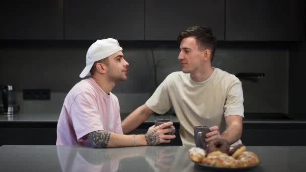 Χαρούμενος Καυκάσιος νεαρός γκέι με τατουάζ που μιλάει με φίλο που φιλάει σύντροφο χαμογελώντας. Χαλαρωμένα θετικά ΛΟΑΤΚΙ ζευγάρι πίνοντας καφέ το πρωί στην κουζίνα στο σπίτι. — Αρχείο Βίντεο