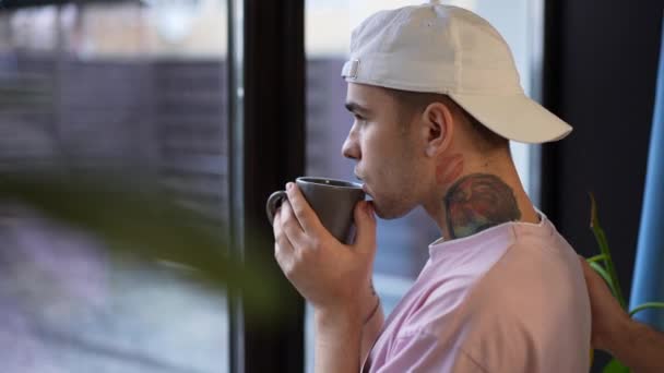 Close-up portrait of thoughtful young gay man looking out window drinking morning coffee as boyfriend entering hugging partner. Happy smiling Caucasian LGBTQ couple enjoying leisure at home. — Stock Video