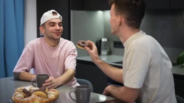 Портрет счастливого белого гея, улыбающегося в качестве партнера, кормящего парня сладким вкусным пончиком. Любимая пара ЛГБТК сидит дома на кухне и наслаждается обедом. — стоковое видео