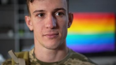 Arka planda gökkuşağı bayrağıyla içeride poz veren eşcinsel askerin portresi. Cesur, genç, beyaz, LGBTQ 'lu adam kendine güvenen bir yüz ifadesiyle kameraya bakıyor..