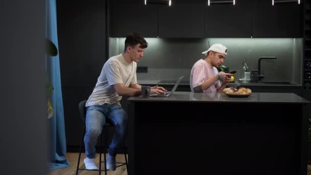Široký záběr mladý kavkazský gayové pár zpráv on-line na notebooku mluvit po telefonu sedí v kuchyni doma. Jistý LGBTQ muži pijí kávu v dopoledních hodinách uvnitř. — Stock video