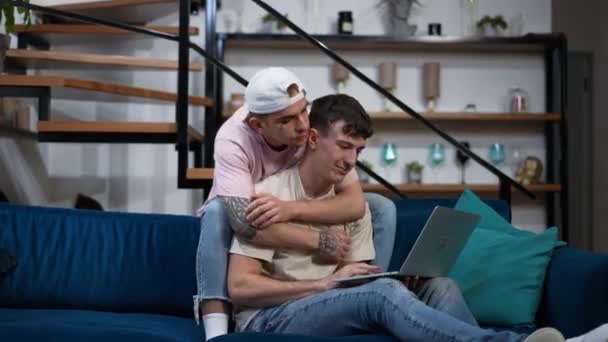 Широкий снимок счастливой белой гей-пары, сидящей на диване в гостиной и занимающейся серфингом в Интернете на ноутбуке. Молодые люди обнимаются в поисках онлайн-фильма. Отдых и отношения на выходных. — стоковое видео