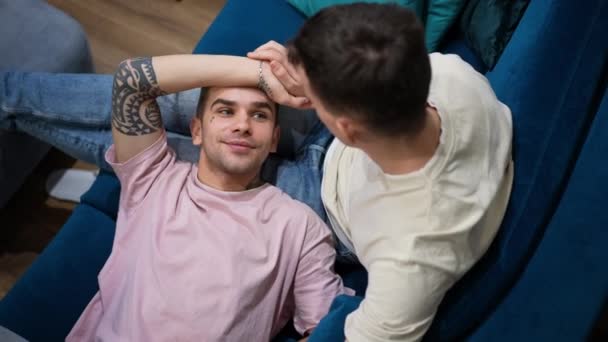Κάτοψη πορτρέτο του χαρούμενου χαλαρού γκέι άντρα που κείτεται στην αγκαλιά του φίλου του χαμογελώντας. Αγαπώντας Καυκάσιος ΛΟΑΤΚΙ ζευγάρι απολαμβάνοντας ελεύθερο χρόνο ανάπαυσης στο σπίτι στον καναπέ στο σαλόνι. — Αρχείο Βίντεο