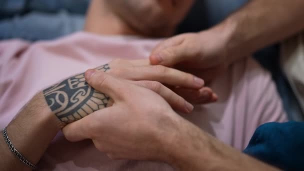 Primer plano tatuado manos con amor irreconocible pareja gay acariciando los dedos. Cámara en vivo zoom hacia fuera como feliz relajado Caucásico LGBTQ pareja acostada en el sofá en casa descansando. — Vídeo de stock