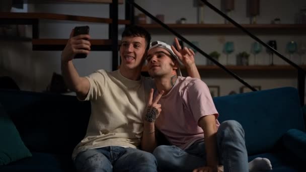 Alegre amante feliz pareja gay haciendo caras gestos de paz tomando selfie en el teléfono inteligente. Hombres LGBTQ caucásicos positivos fotografiando en la sala de estar en casa haciendo muecas. — Vídeo de stock