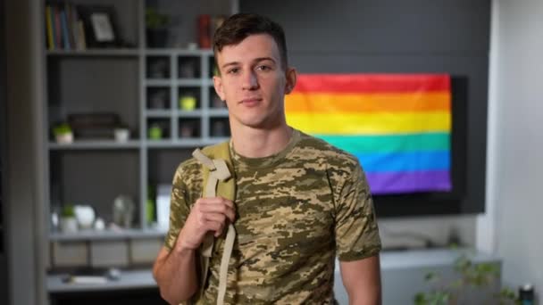 Porträtt av självsäker ung man i kamouflage T-shirt tittar på kameran med regnbåge hbtq flagga i bakgrunden. Modig vit gay soldat poserar inomhus hemma. — Stockvideo