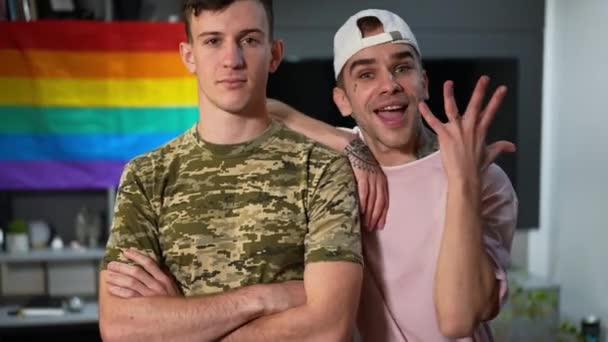 Ritratto di giovane gay eccitato che vanta anello di fidanzamento in piedi con fidanzato militare al chiuso. Felice caucasico LGBTQ coppia guardando fotocamera vantarsi proposta con bandiera arcobaleno sullo sfondo. — Video Stock