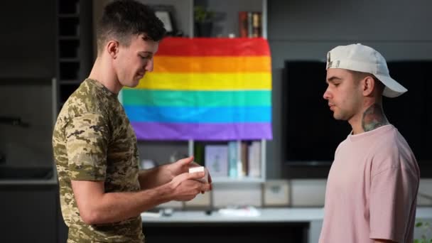 Vista lateral joven gay hombre negando propuesta de militar socio en el interior de casa. Caucásico tatuado LGBTQ persona dejando triste novio solo en el interior. Bandera del arco iris colgando de fondo. — Vídeos de Stock