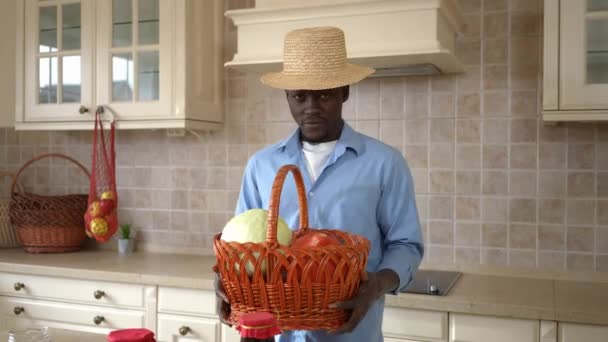 Afroamerykanin młody rolnik w słomkowym kapeluszu rozciągający kosz ze żniwami uśmiechnięty patrząc w kamerę. Portret zadowolonego mężczyzny pozującego w kuchni z organicznymi warzywami. — Wideo stockowe