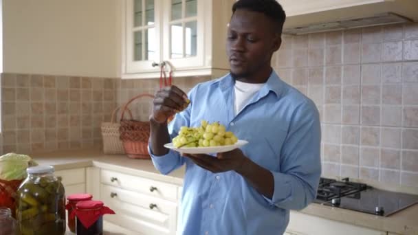 Молодой афроамериканец пробует вкусный виноград, стоя дома на кухне. Портрет уверенного фермера, поедающего вкусные спелые фрукты в помещении. Садоводство и здоровое питание. — стоковое видео
