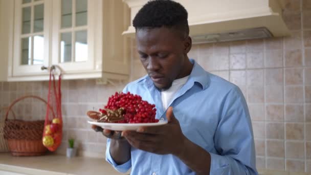 Um jovem afro-americano a cheirar bagas vermelhas de viburnum a fazer expressão facial nojenta. Retrato de cara com colheita madura em pé na cozinha em casa dentro de casa. — Vídeo de Stock